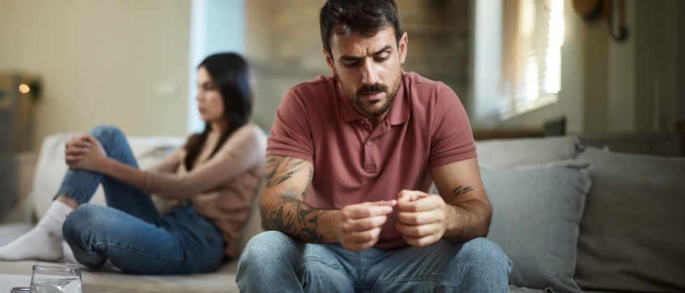 Man is sitting with his back to a woman, looking insecurely at his fingers.
